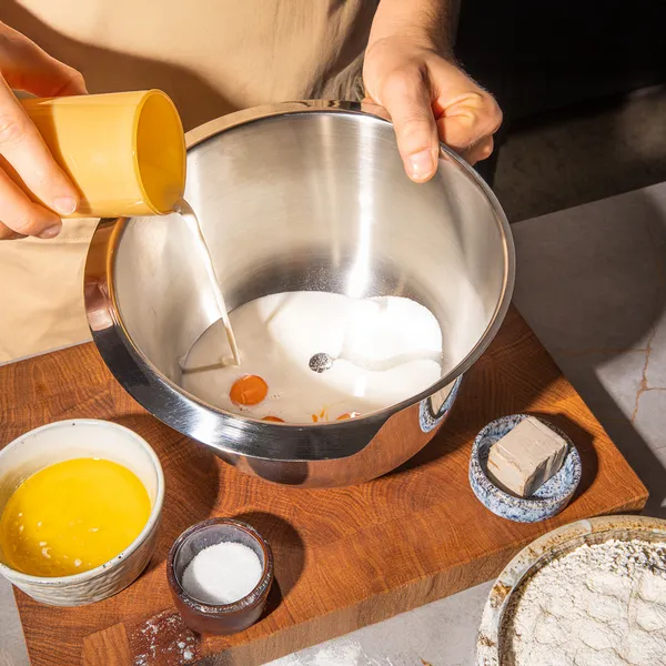 Hands mixing ingredients in a metal bowl on a wooden board, with eggs, flour, and other baking supplies visible.
