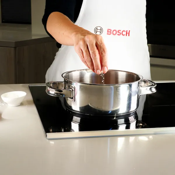 A person in a Bosch apron sprinkles ingredients into a pot on an induction cooktop.