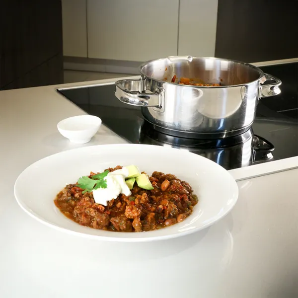 A bowl of chili with avocado and sour cream, with a pot of chili on an induction cooktop in the background.