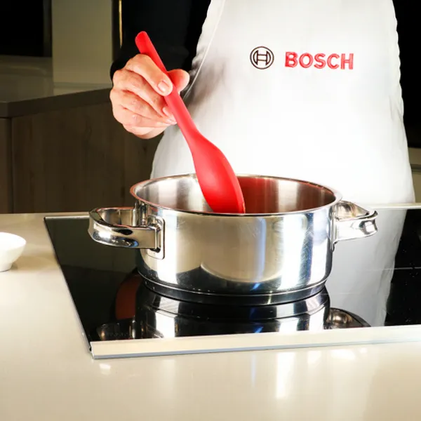Person wearing a Bosch apron stirring a stainless steel pot with a red silicone spatula on a stove.