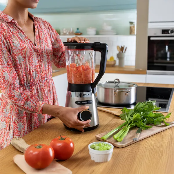 Person using a Bosch blender in a kitchen, blending chopped tomatoes besides other vegetables on a wooden countertop.