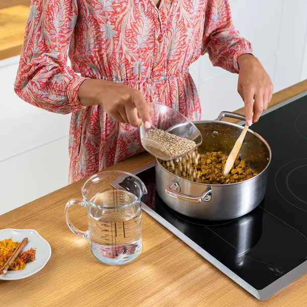 Person in patterned dress pouring grains into a pot while stirring on a kitchen stovetop.