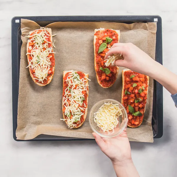 Four French bread halves on a baking tray lined with parchment paper topped with tomato sauce and basil leaves with a person sprinkling shredded cheese on one half.