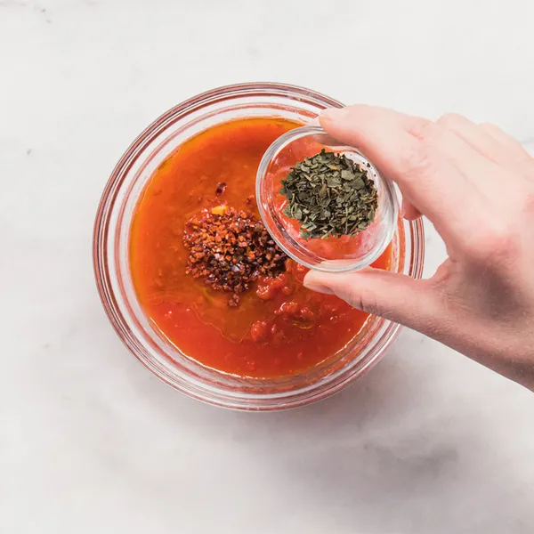 Hand holding a small glass bowl with dried herbs above a glass bowl containing red sauce with chili flakes on a white surface.