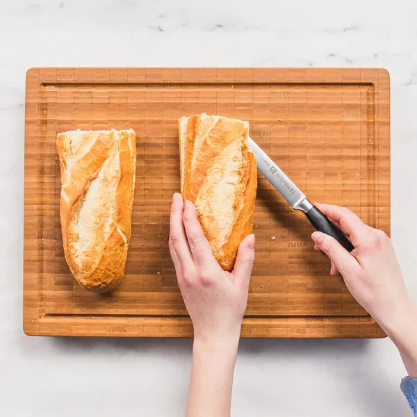 Two small loaves of French bread on a wooden cutting board with a hand holding one loaf and a knife poised to cut it.