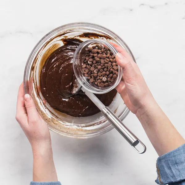 Hands in white apron pouring vanilla into a glass bowl with eggs and chocolate batter, red spatula on striped cloth