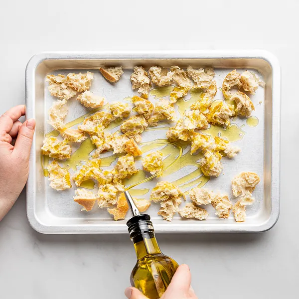 Hands drizzling olive oil over torn pieces of bread on a baking sheet lined with parchment paper.