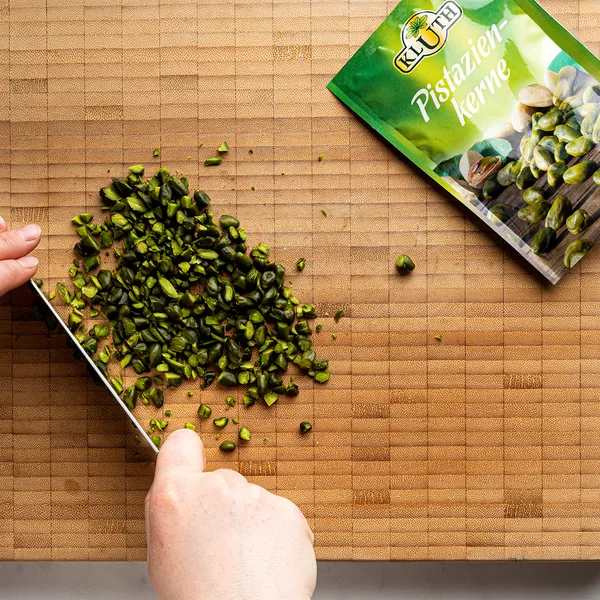 Hands chopping green pistachio kernels on a cutting board next to a package of Kluth pistachio kernels.