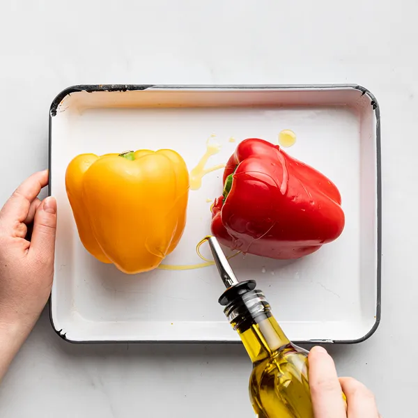 Two bell peppers, one yellow and one red, on a tray being drizzled with olive oil from a bottle.