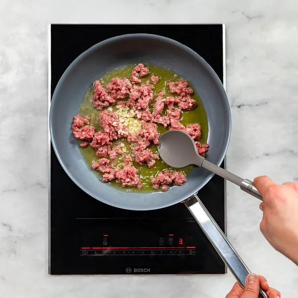 Sausages cooking in a frying pan with oil and minced garlic on an induction stove with a hand stirring with a spoon.