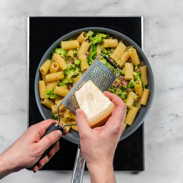 Hands grating a block of hard cheese over a pan of rigatoni pasta with broccoli and bits of meat.