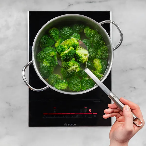 Hand holding a spoon stirring broccoli florets boiling in a pot on a Bosch induction cooktop.