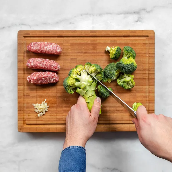 Hands cutting broccoli into florets on a wooden cutting board with minced garlic and three raw sausage links nearby.