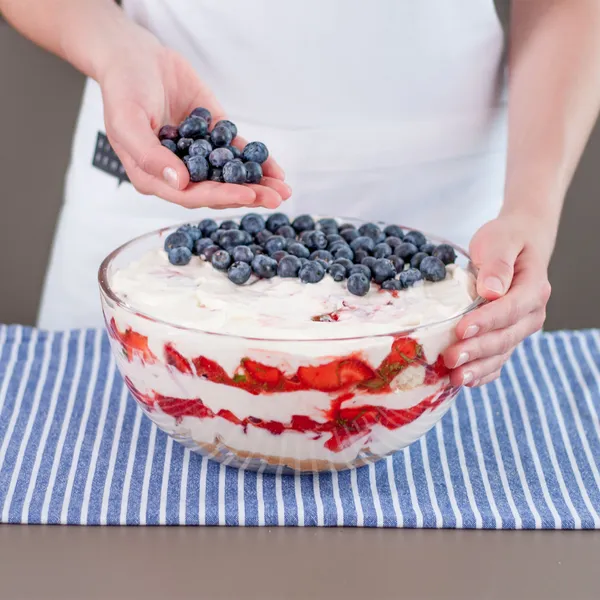 Close-up of a person's hands in a white apron sprinkling fresh blueberries onto a large glass bowl of layered strawberry-and-cream trifle, set on a blue-and-white striped cloth on a kitchen counter.