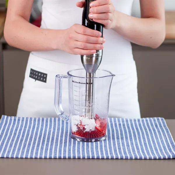 Hands of a person in a white apron holding a black immersion blender inside a clear measuring jug with sliced strawberries and white powder, set on a blue-and-white striped cloth on a kitchen counter.
