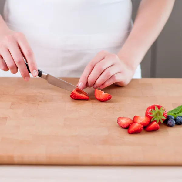 Close-up of a person's hands slicing a strawberry with a kitchen knife on a wooden cutting board, with a small pile of sliced strawberries, a whole strawberry, blueberries, and mint nearby.
