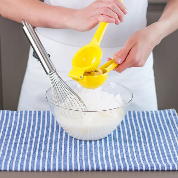 Hands of a person in a white apron squeezing a yellow handheld citrus juicer over a glass mixing bowl containing a white powder and a metal whisk, set on a blue-and-white striped cloth on a kitchen counter.