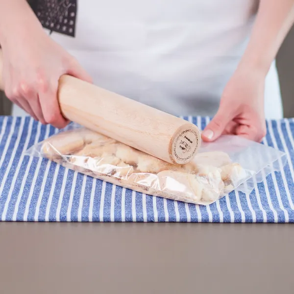 Person crushing cookies in a plastic bag using a wooden rolling pin on a blue-striped cloth.