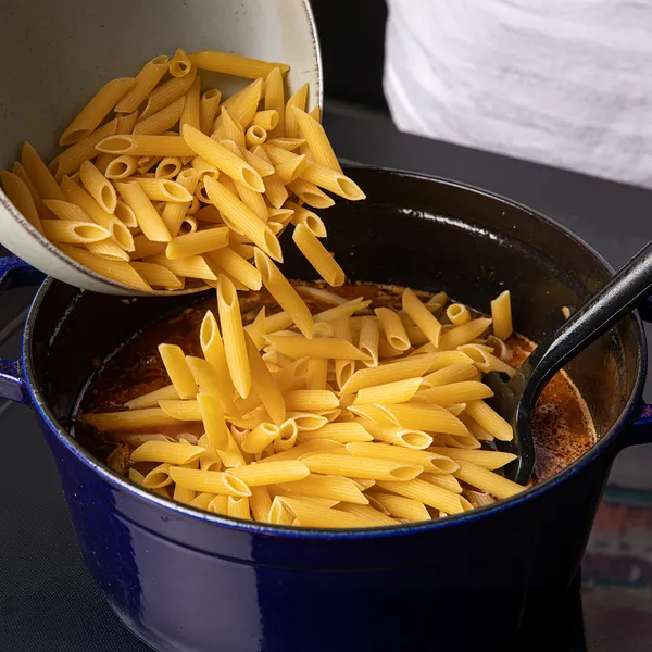 Uncooked penne pasta being poured from a bowl into a large blue pot with sauce and a black spoon inside.