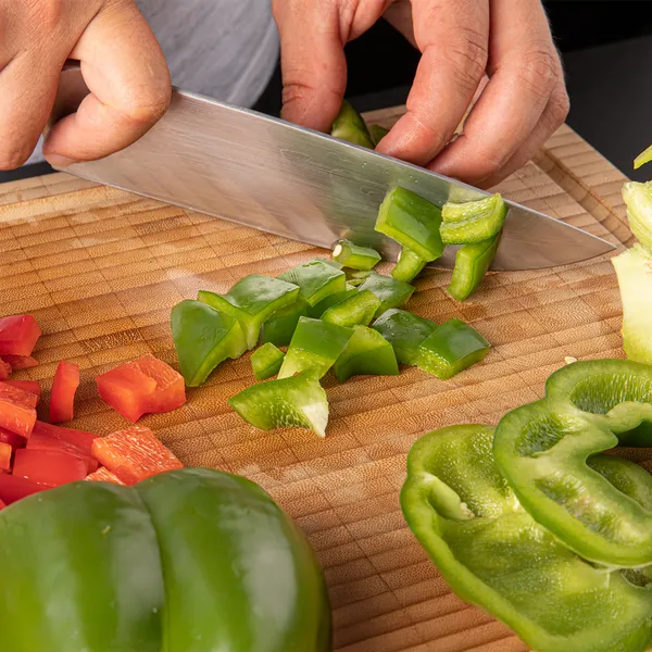 Hands chopping green bell pepper on a wooden cutting board with sliced red and green bell peppers nearby.