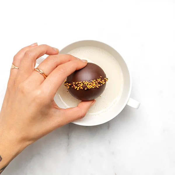 Overhead view of a placing a chocolate-coated cocoa bomb with orange sprinkles into a white mug of milk on a marble surface.