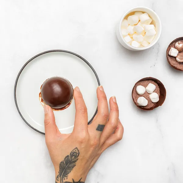 Overhead view of a hand placing a glossy chocolate cocoa bomb on a small saucer, with two open chocolate shells topped with mini marshmallows and a bowl of marshmallows on a white marble surface.