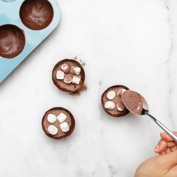 Overhead view of a hand spooning cocoa mixture into chocolate half-shells topped with mini marshmallows, with a blue silicone mold visible on a white marble surface.