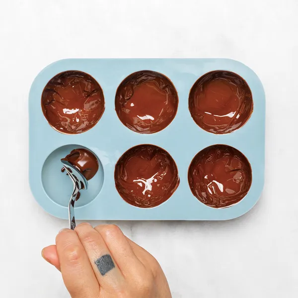 Overhead view of a hand spooning melted chocolate into a blue six-cavity silicone half-sphere mold on a white surface.