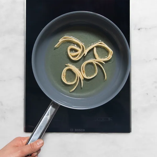 Hand holding frying pan with swirled batter cooking on a black stovetop, white marble counter background.
