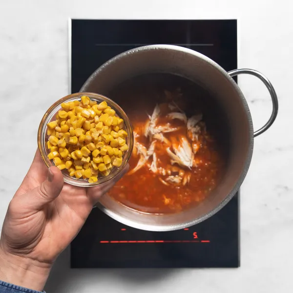 Hand adding a small bowl of corn to a pot with shredded chicken and red sauce on a stovetop.