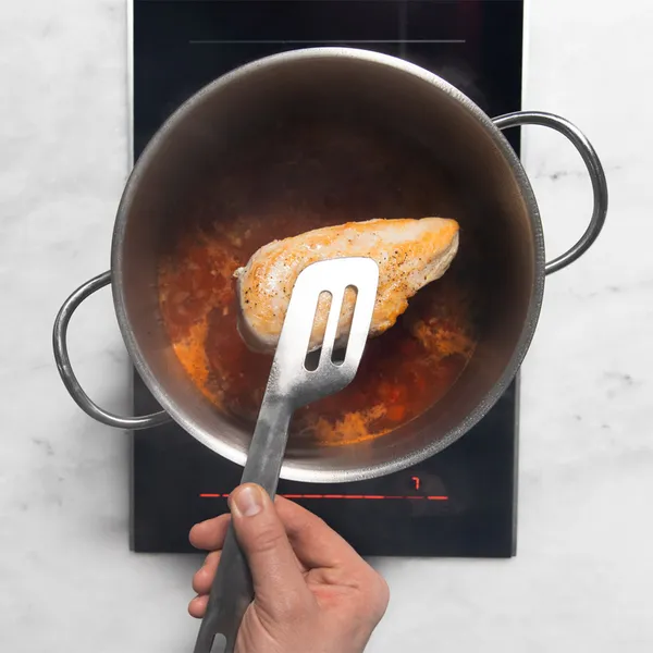 Hand cooking a chicken breast in a pot with broth on an induction cooktop, viewed from above.