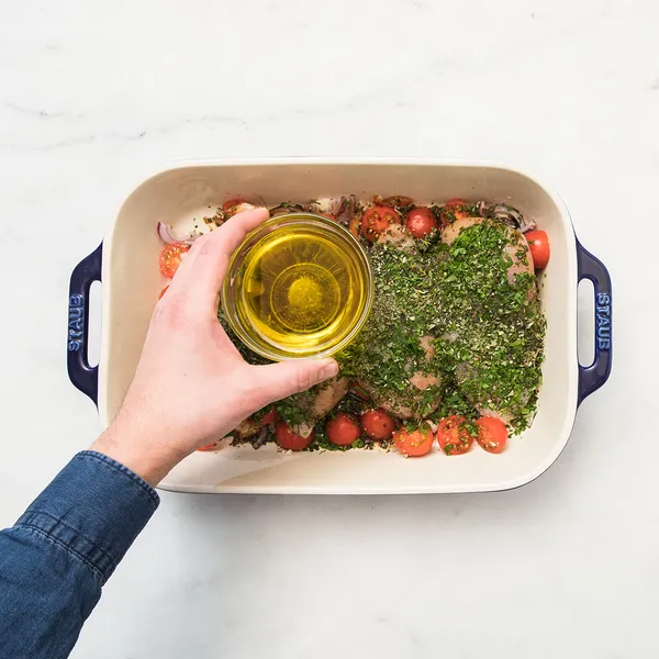 Hand pouring oil over herb-seasoned chicken and cherry tomatoes in a baking dish on a white marble surface.
