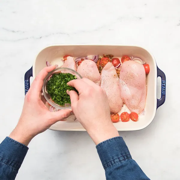 Hands sprinkling chopped herbs over raw chicken breasts, cherry tomatoes, and onions in a baking dish on a marble surface.