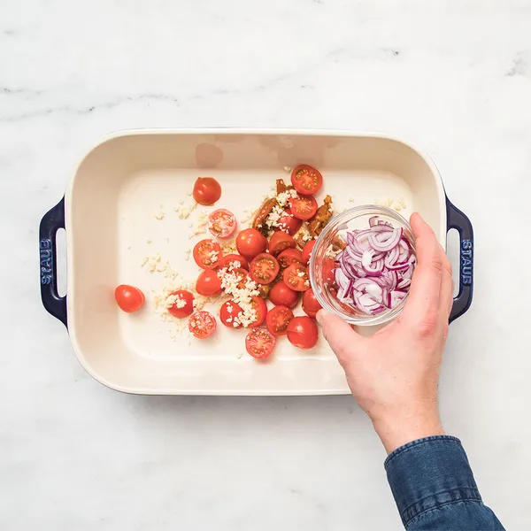 Hand adding sliced red onions to a baking dish with halved cherry tomatoes and minced garlic on a white marble surface.