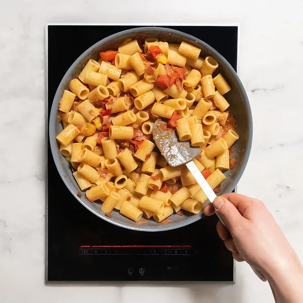 Hand stirring rigatoni pasta with diced tomatoes in a frying pan on an induction cooktop.