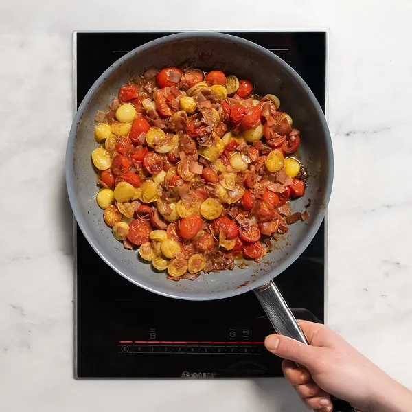 Hand holding a frying pan with bacon and cherry tomatoes on an induction cooktop.