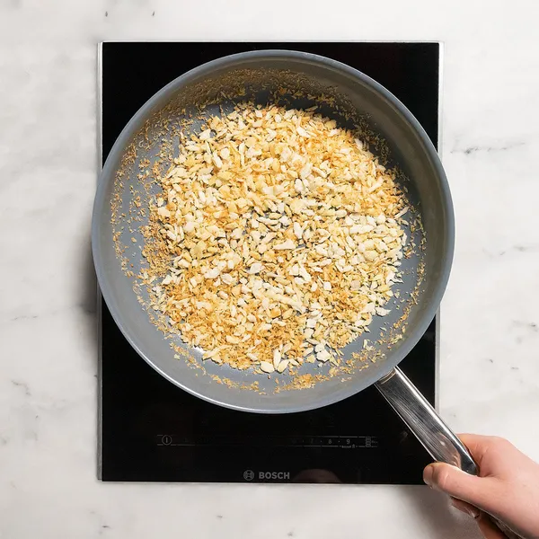 Hand holding a frying pan with toasted panko breadcrumbs on an induction cooktop.