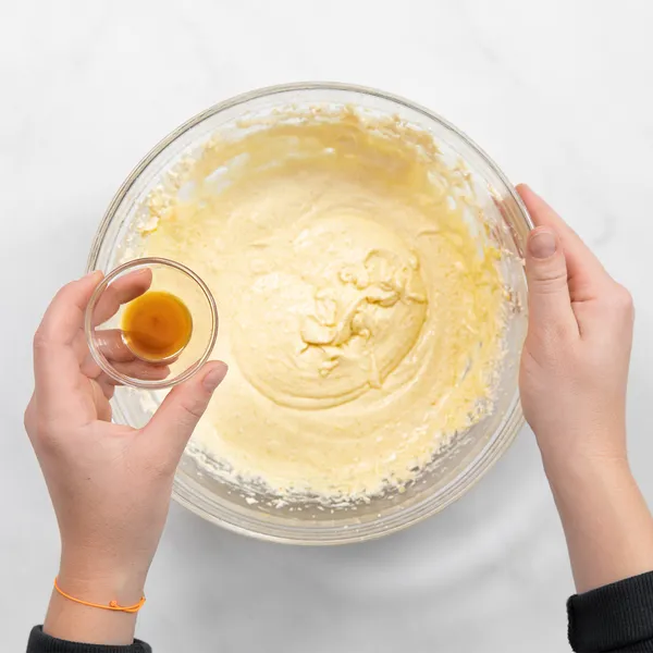 Hands holding a small bowl of vanilla extract over a glass bowl of apple cider donut batter, ready to add the ingredient