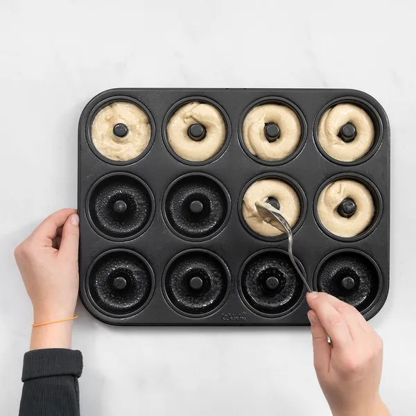 Hands dipping a donut batter‑filled donut pan into the oven, ready to bake apple cider donuts