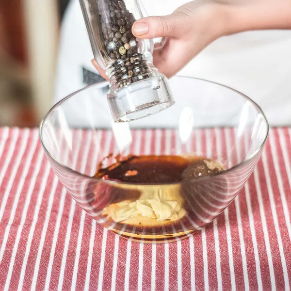 A hand holds a clear pepper grinder, adding black peppercorns to a glass bowl containing various sauces and ingredients on a red and white striped tablecloth.