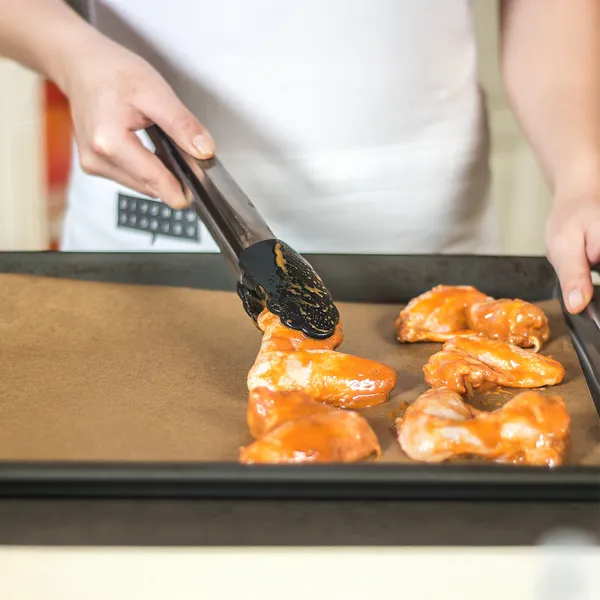 A person in a white apron uses tongs to place raw, sauce-covered chicken wings onto a baking sheet lined with parchment paper.