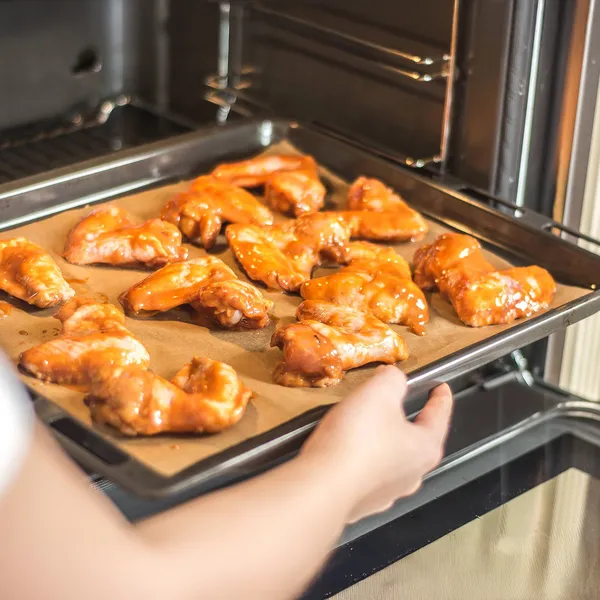 Person placing a baking tray with glazed chicken wings into an oven lined with parchment paper.