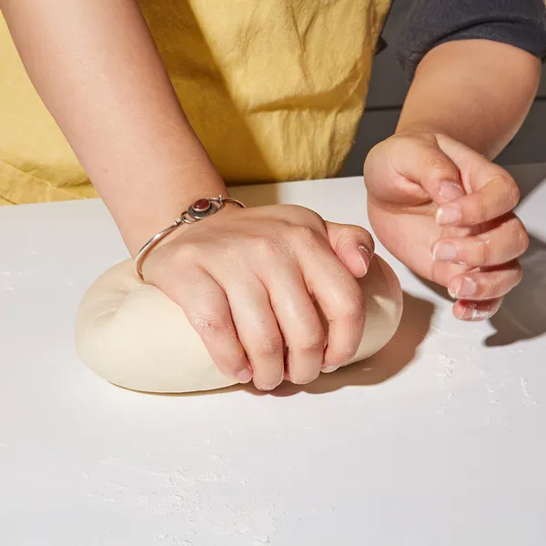 Hands kneading dough on a white surface, with a person wearing a yellow apron and a silver bracelet.