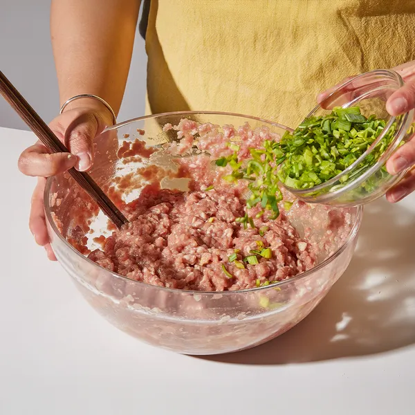 Person adding chopped green onions to ground meat in a glass bowl, mixing with chopsticks on a white countertop.