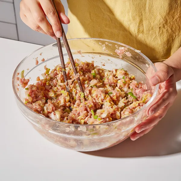 Person mixing ground meat and vegetables in a glass bowl using chopsticks on a white countertop.