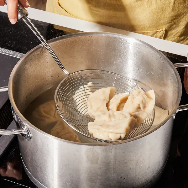 Hands using a wire skimmer to lift boiled dumplings from a stainless-steel pot on a stovetop.