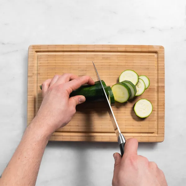 Hands slicing a zucchini on a wooden cutting board with a kitchen knife.