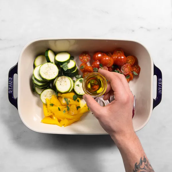Hand holding a small glass container of oil above a baking dish with sliced zucchini, halved cherry tomatoes, and yellow bell pepper pieces.