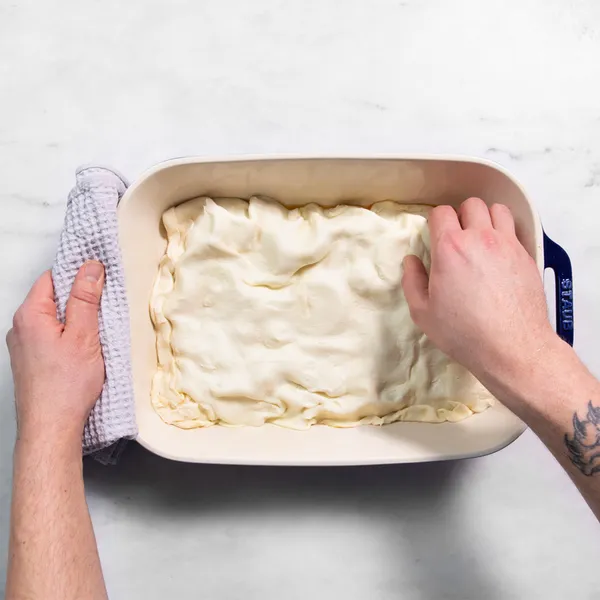 Hands pressing dough into a rectangular baking dish on a white surface.