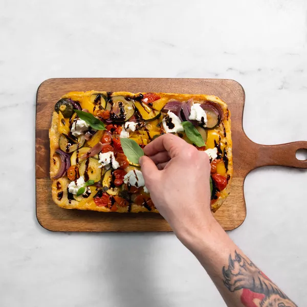 Hand with tattoo placing fresh basil leaves on rectangular veggie pizza with cherry tomatoes, red onions, and dollops of cheese on wooden board.